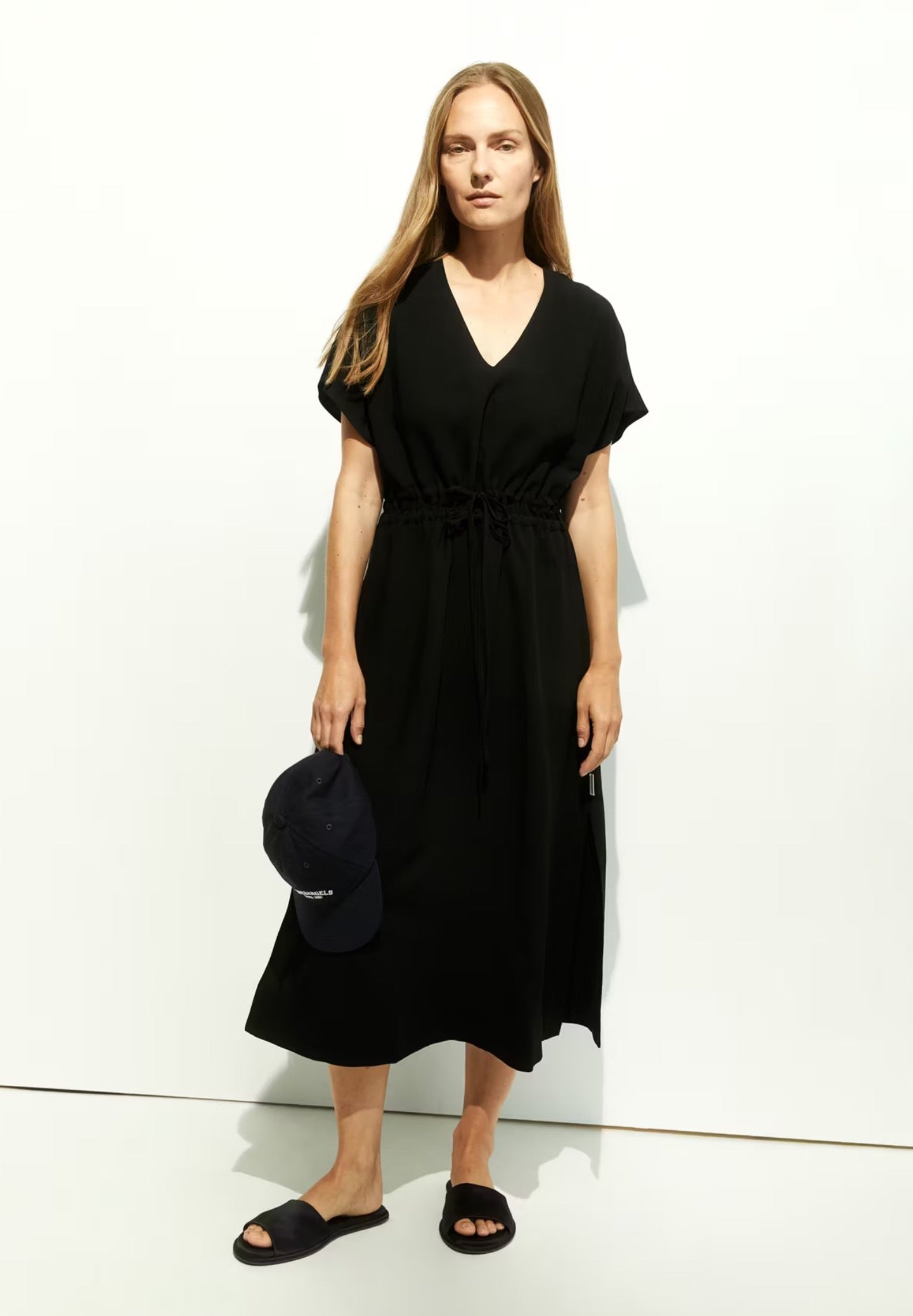 A woman models a black LENZING™ ECOVERO™ and linen blend dress from Armedangels. The dress features a deep V-neckline, short sleeves with a slight flare, and a drawstring waist that creates a cinched silhouette. The hemline reaches just below the knees, and the lightweight, flowy fabric drapes naturally. The person holds a black cap in one hand, wears black slide sandals, and has long, straight hair. They stand against a plain white background with a subtle shadow cast to the side.