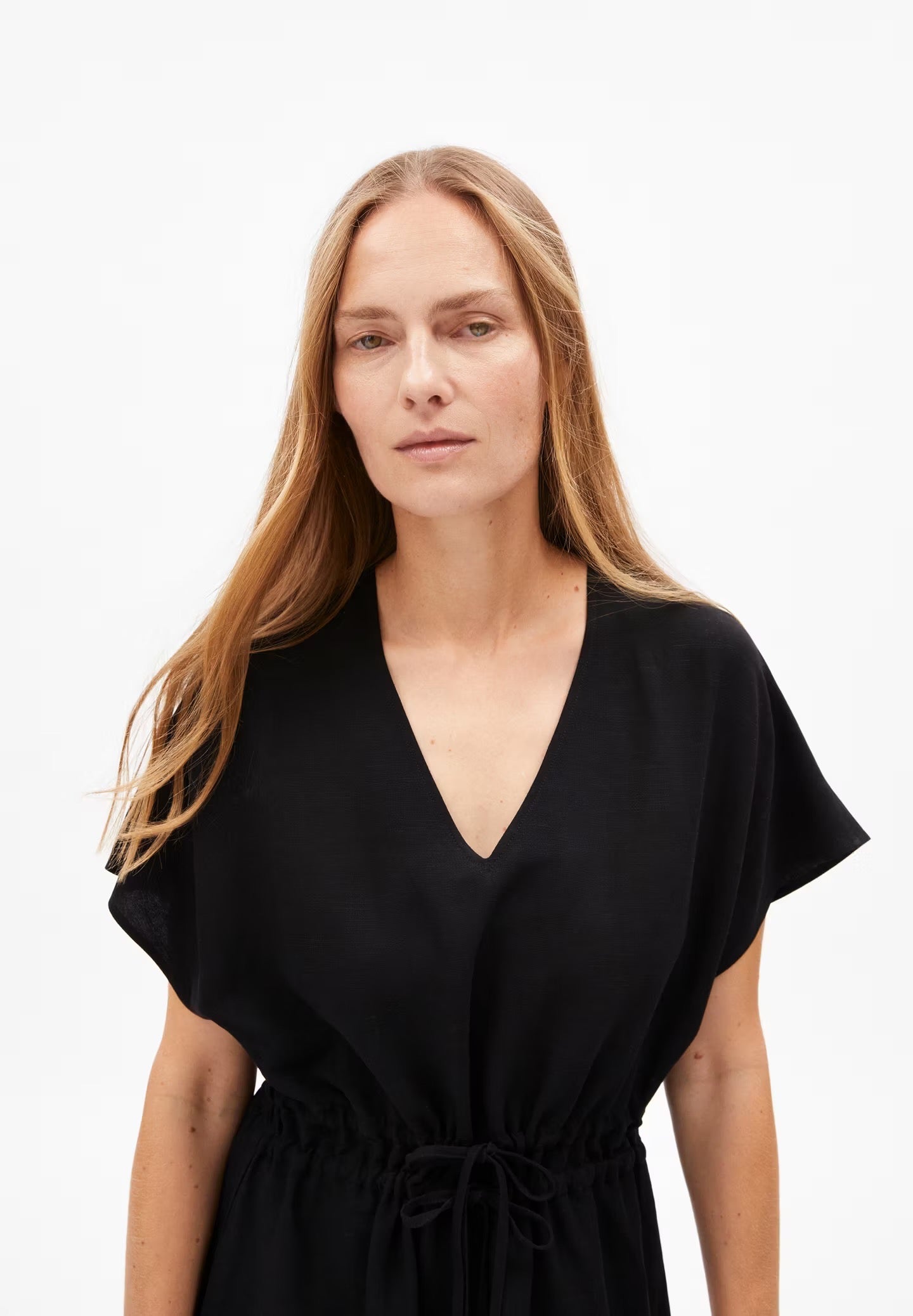 A woman models a black LENZING™ ECOVERO™ and linen blend dress from Armedangels. The dress features a deep V-neckline, short sleeves with a slight flare, and a drawstring waist that creates a cinched silhouette. The fabric appears lightweight and flowy, with a minimalist design, set against a plain white background. The person has long, straight hair and stands with one hand on their hip.
