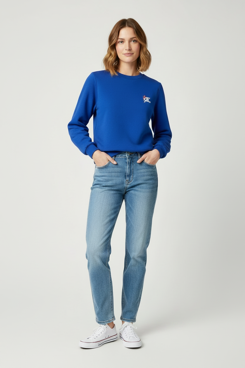A woman modeling a royal blue crewneck sweatshirt with a small embroidered logo on the left chest, paired with light-wash blue jeans and white sneakers, against a light gray studio background.
