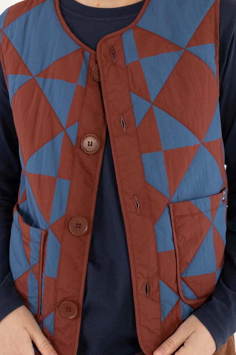 Quilted thermo gilet from Danefae Copenhagen. Regular length, round neck, button closure with four buttons, roomy patch pockets. Featuring a geometric pattern of diametrically placed squares and triangles in brick red and denim blue. Made from nylon.