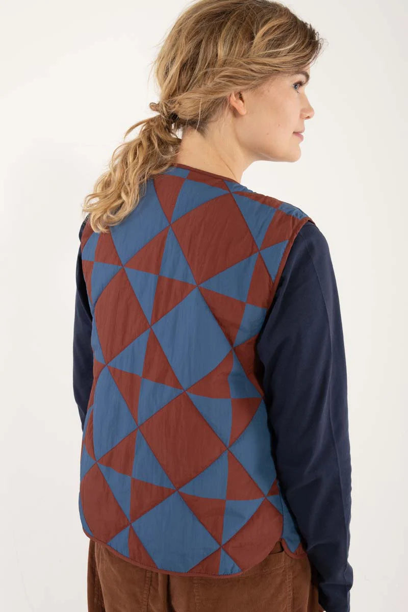 Quilted thermo gilet from Danefae Copenhagen. Regular length, round neck, button closure with four buttons, roomy patch pockets. Featuring a geometric pattern of diametrically placed squares and triangles in brick red and denim blue. Made from nylon.