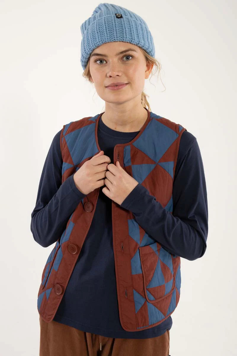 Quilted thermo gilet from Danefae Copenhagen. Regular length, round neck, button closure with four buttons, roomy patch pockets. Featuring a geometric pattern of diametrically placed squares and triangles in brick red and denim blue. Made from nylon.