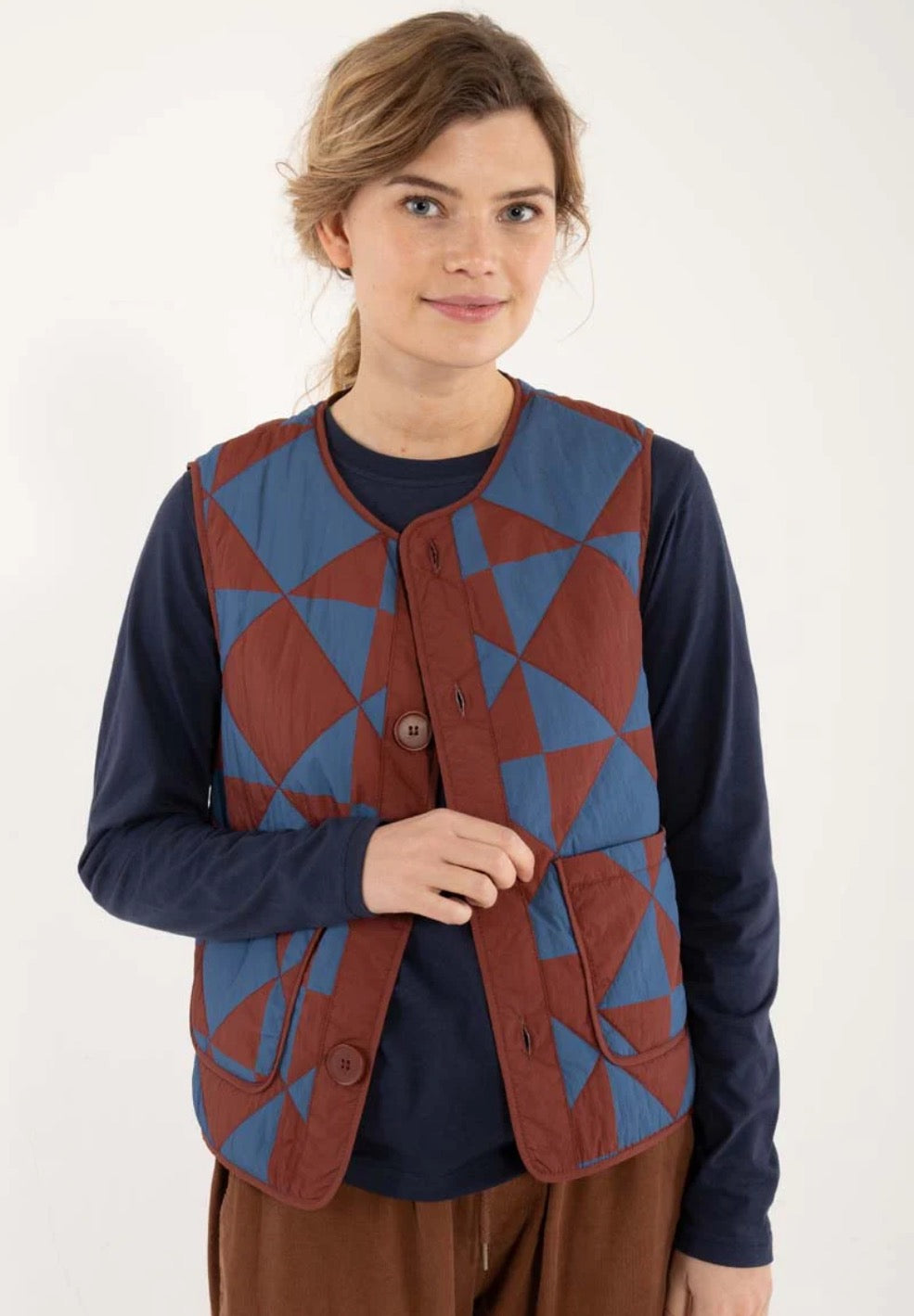 Quilted thermo gilet from Danefae Copenhagen. Regular length, round neck, button closure with four buttons, roomy patch pockets. Featuring a geometric pattern of diametrically placed squares and triangles in brick red and denim blue. Made from nylon.