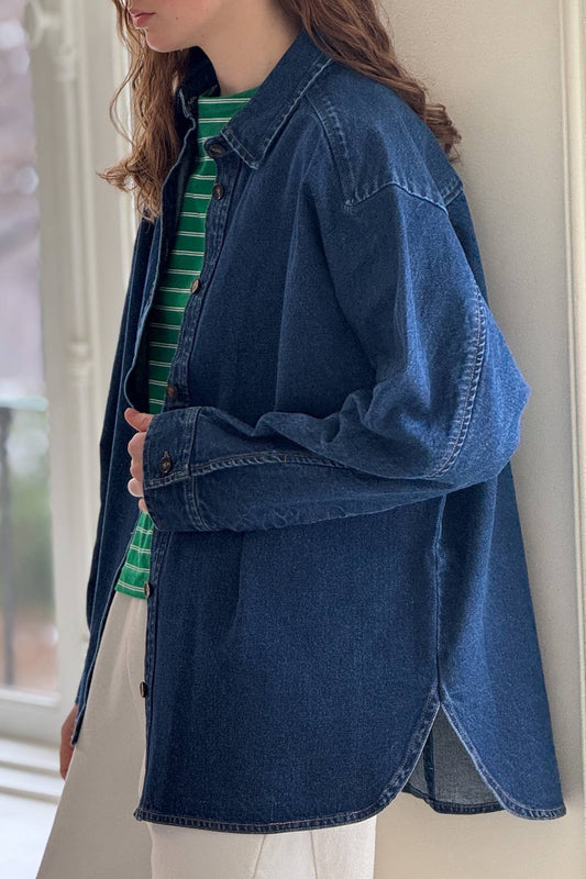 A woman with long, wavy hair stands near a window, wearing a dark blue cotton denim shirt from Le Bon Shoppe. The shirt features a button-down front, a small collar, and long sleeves with buttoned cuffs, layered over a green and white striped shirt, and paired with relaxed white trousers. The slightly oversized denim shirt creates a casual, effortless look, with natural light from the window softly illuminating the scene.