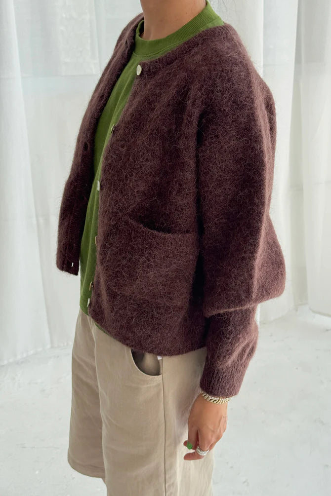 Person wearing a brown textured Amelie cardigan from Le Bon Shoppe over a green shirt with beige pants against a white curtain background