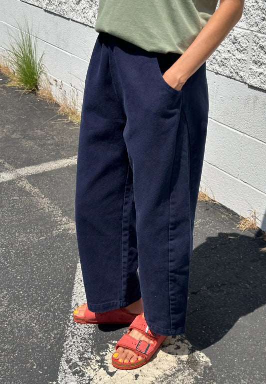 Arc Pants from Le Bon Shoppe. Barrel leg trousers made from durable cotton canvas. Featuring an elastic high waist, deep side pockets, and a slightly curved, cropped leg with a unique side panel. In a dark navy colour.