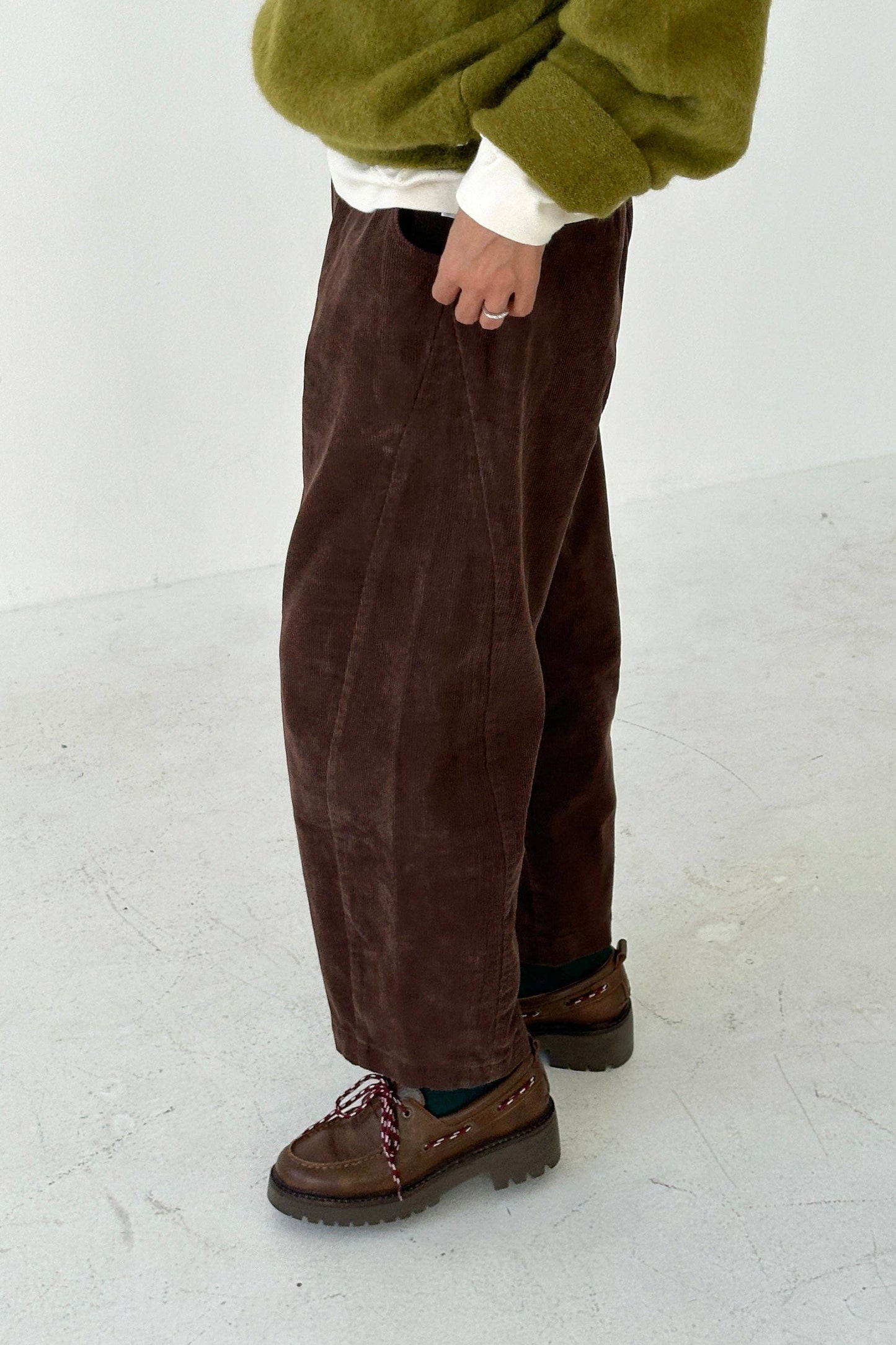 Person wearing chocolate brown corduroy barrel-leg Arc pants from Le Bon Shoppe and green sweater on a white background