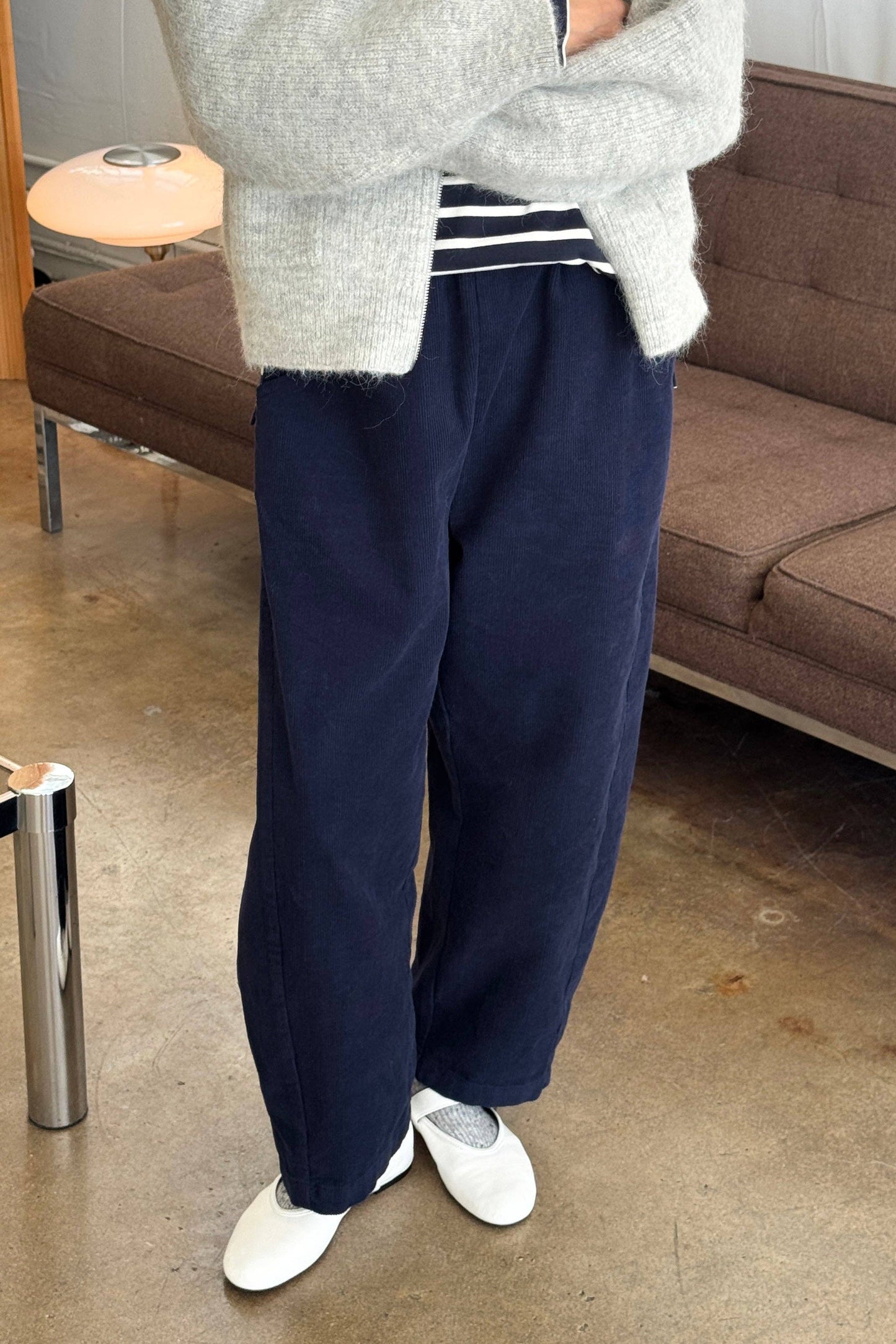 Person wearing a gray sweater, navy corduroy barrel leg Arc pants from Le Bon Shoppe, and white shoes in a living room setting.