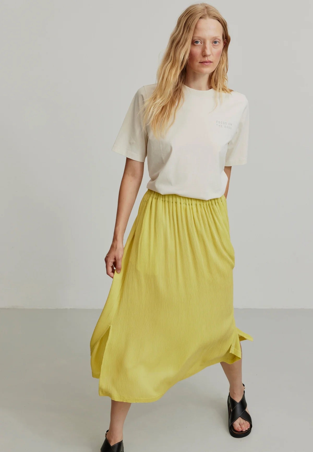 A person models a vibrant mustard yellow LENZING™ ECOVERO™ midi skirt from Recolution, featuring a high-waisted, elastic waistband with a gathered design and a subtle side slit. The skirt has a soft, crinkled texture and flows gracefully, paired with a white short-sleeve t-shirt and black strappy sandals. The person stands against a plain light gray background, with one hand in the skirt's side pocket and the other resting by their side.