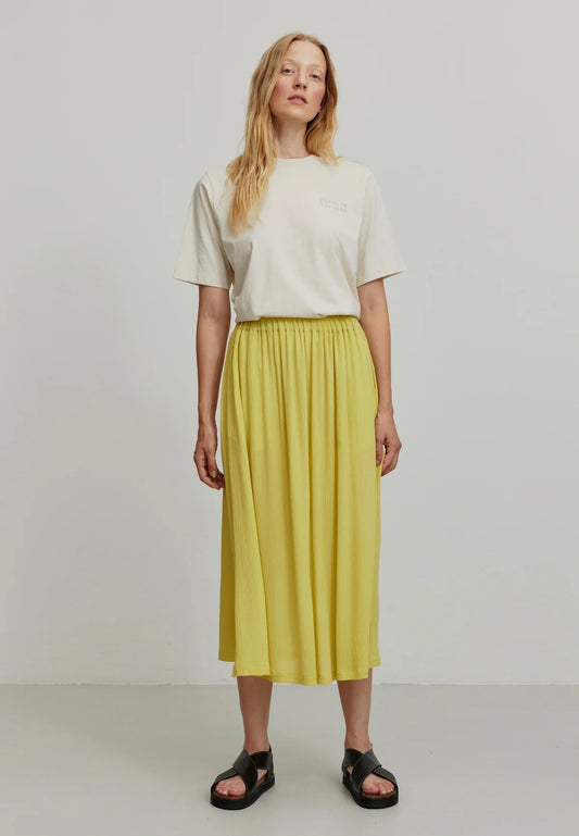 A person models a vibrant mustard yellow LENZING™ ECOVERO™ midi skirt from Recolution, featuring a high-waisted, elastic waistband with a gathered design and a subtle side slit. The skirt has a soft, crinkled texture and flows gracefully, paired with a white short-sleeve t-shirt and black strappy sandals. The person stands against a plain light gray background, with one hand in the skirt's side pocket and the other resting by their side.