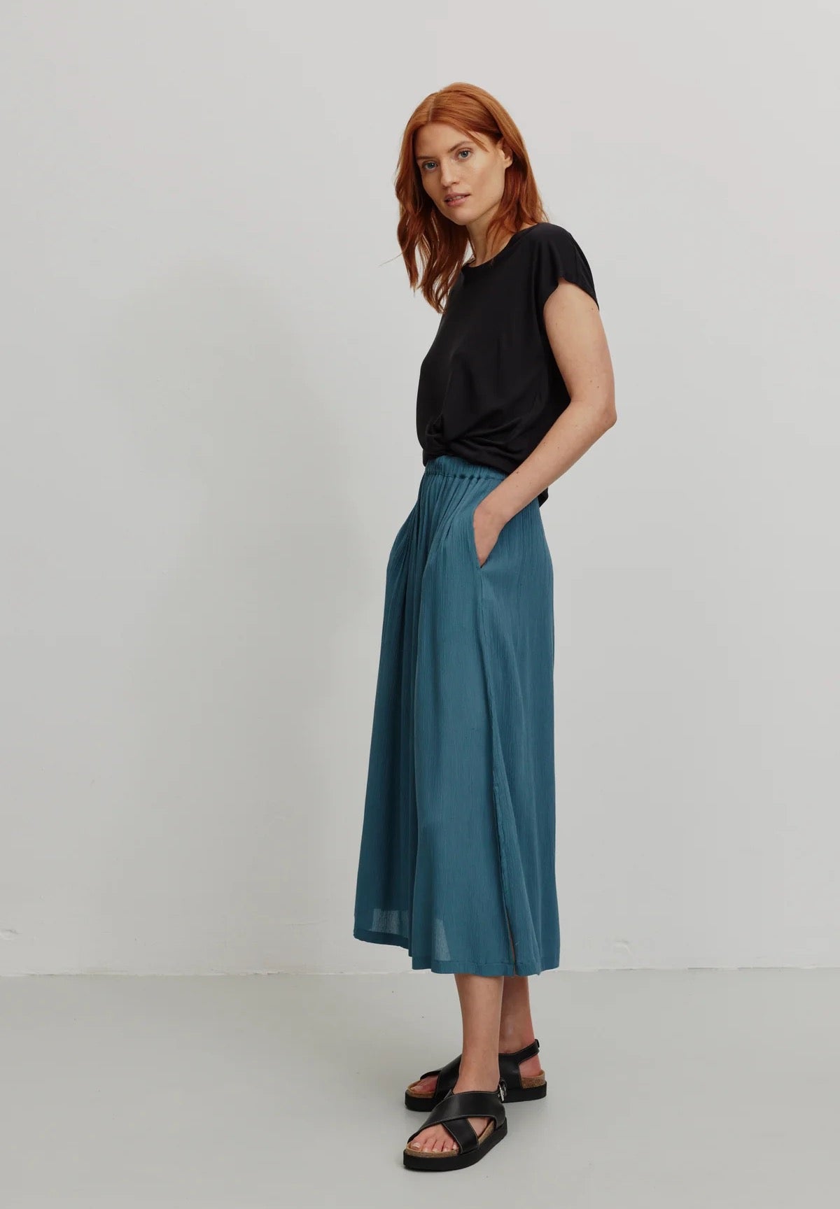 A person models a rich teal LENZING™ ECOVERO™ midi skirt from Recolution, featuring a high-waisted, elastic waistband with a gathered design and a subtle front slit. The skirt has a soft, crinkled texture and flows gracefully, paired with a black short-sleeve t-shirt and black strappy sandals. The person stands against a plain light gray background, with one hand in the skirt's side pocket and the other resting by their side, showcasing the skirt's elegant silhouette.