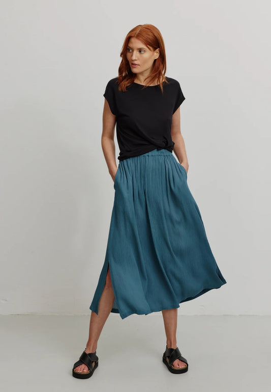 A person models a rich teal LENZING™ ECOVERO™ midi skirt from Recolution, featuring a high-waisted, elastic waistband with a gathered design and a subtle front slit. The skirt has a soft, crinkled texture and flows gracefully, paired with a black short-sleeve t-shirt and black strappy sandals. The person stands against a plain light gray background, with one hand in the skirt's side pocket and the other resting by their side, showcasing the skirt's elegant silhouette.