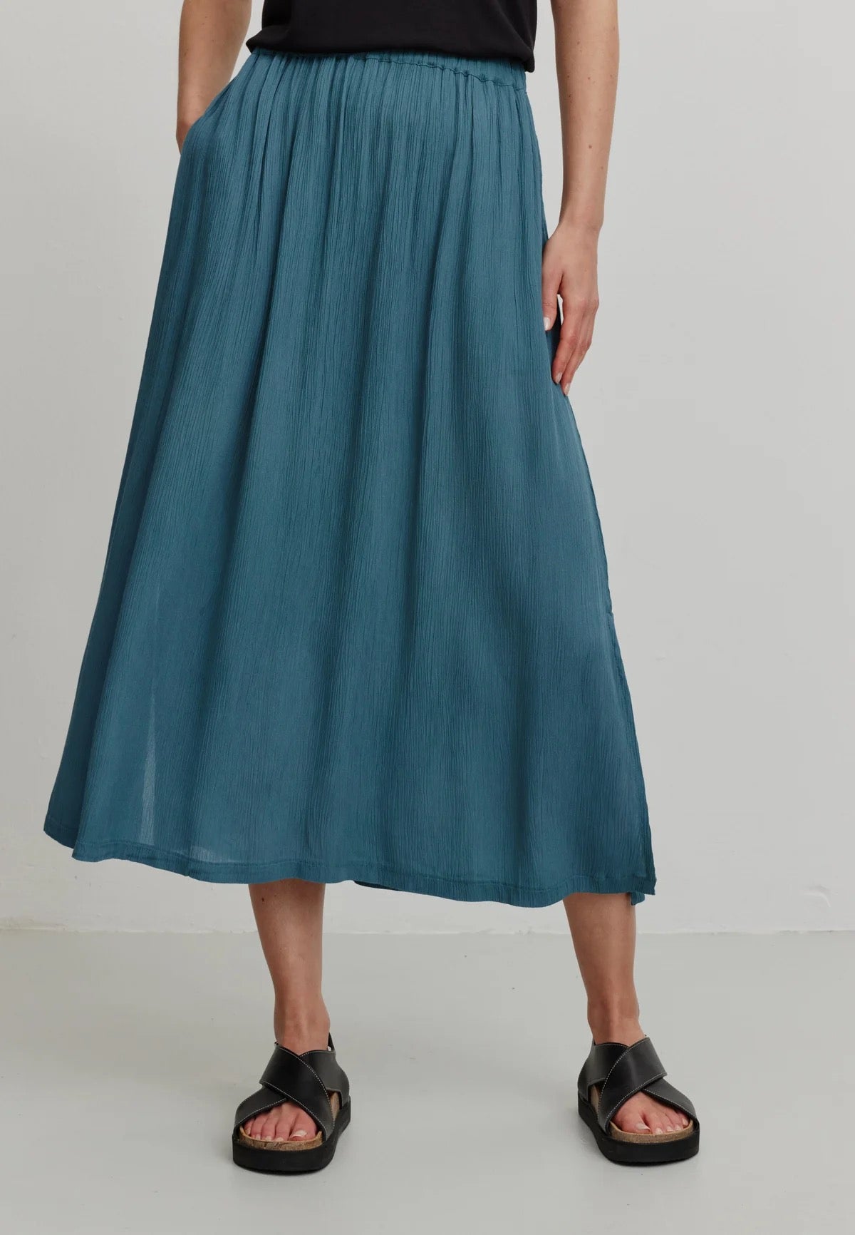 A person models a rich teal LENZING™ ECOVERO™ midi skirt from Recolution, featuring a high-waisted, elastic waistband with a gathered design and a subtle front slit. The skirt has a soft, crinkled texture and flows gracefully, paired with a black short-sleeve t-shirt and black strappy sandals. The person stands against a plain light gray background, with one hand in the skirt's side pocket and the other resting by their side, showcasing the skirt's elegant silhouette.