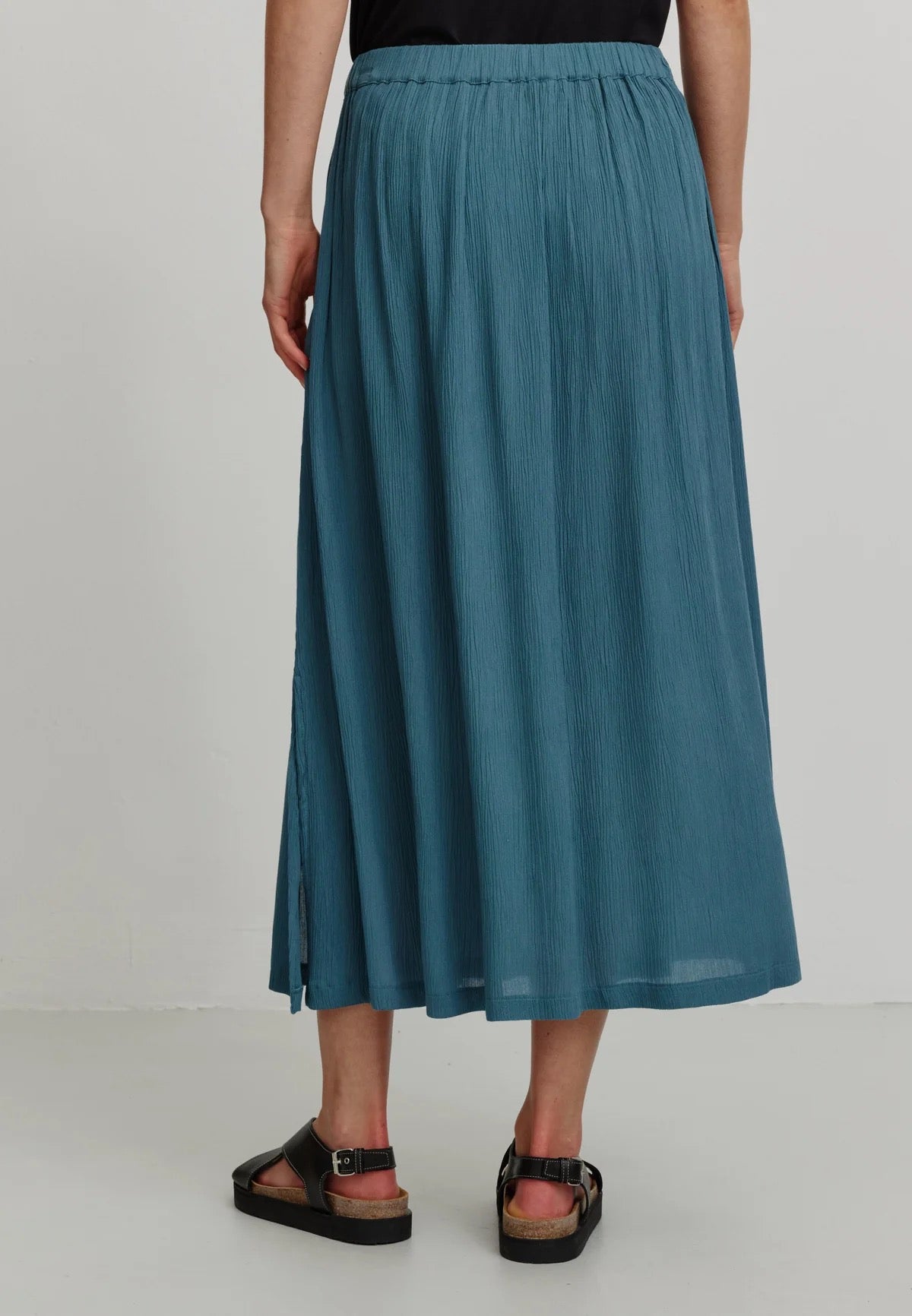 A person models a rich teal LENZING™ ECOVERO™ midi skirt from Recolution, featuring a high-waisted, elastic waistband with a gathered design and a subtle front slit. The skirt has a soft, crinkled texture and flows gracefully, paired with a black short-sleeve t-shirt and black strappy sandals. The person stands against a plain light gray background, with one hand in the skirt's side pocket and the other resting by their side, showcasing the skirt's elegant silhouette.