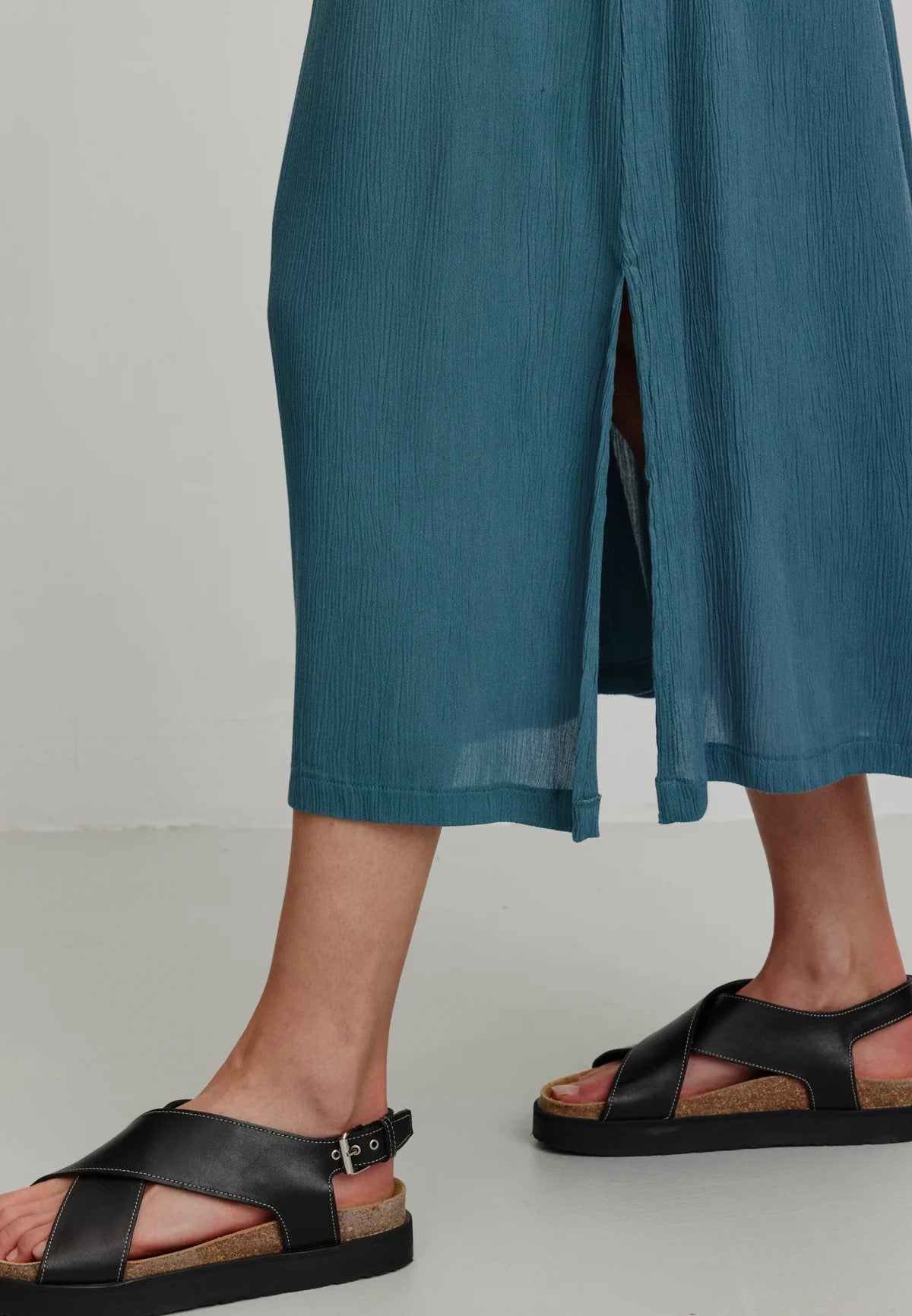 A person models a rich teal LENZING™ ECOVERO™ midi skirt from Recolution, featuring a high-waisted, elastic waistband with a gathered design and a subtle front slit. The skirt has a soft, crinkled texture and flows gracefully, paired with a black short-sleeve t-shirt and black strappy sandals. The person stands against a plain light gray background, with one hand in the skirt's side pocket and the other resting by their side, showcasing the skirt's elegant silhouette.