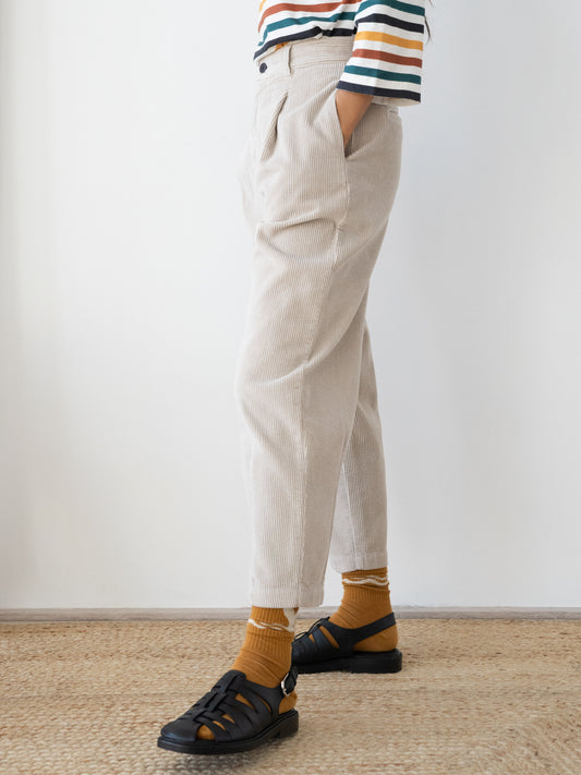 A person wearing light beige Haruku corduroy trousers from TWOTHIRDS, mustard yellow socks, and black sandals, standing on a woven jute rug against a plain white wall.