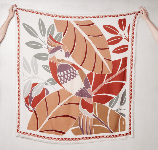 Women’s scarf from the French fashion brand Maradji. Tropical leaves and a parrot in shades terracotta, sage and caramel on a oat milk coloured background. Square shape, made from a sustainable wool and silk blend.