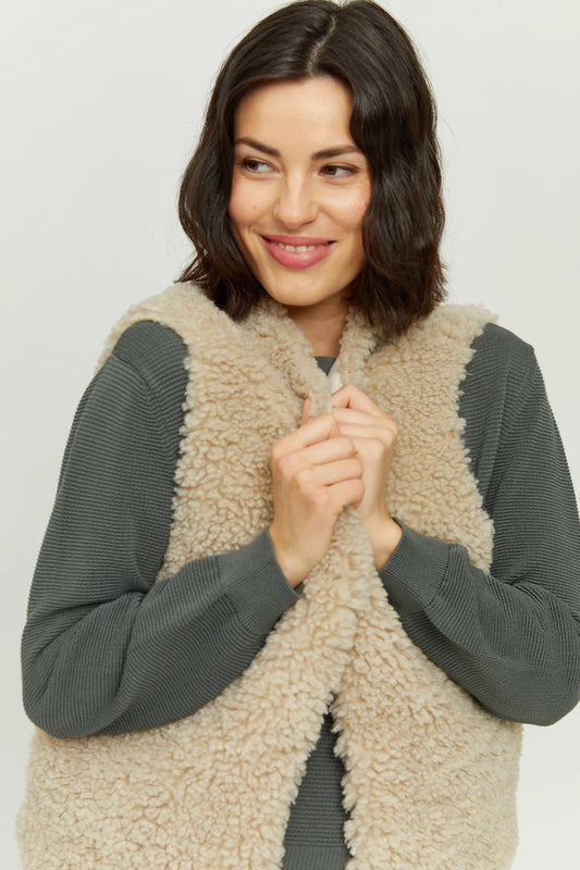 A woman wearing a sand coloured teddy fleece gilet by Mazine, over a sage coloured long sleeve top.