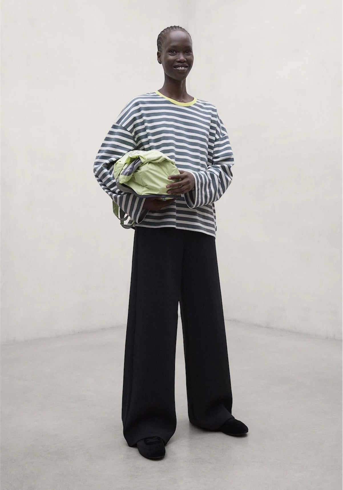 A model stands in a minimalist studio setting wearing a sage and off white striped recycled cotton long-sleeve top by Ecoalf black wide-leg trousers, and holding a light green bag.