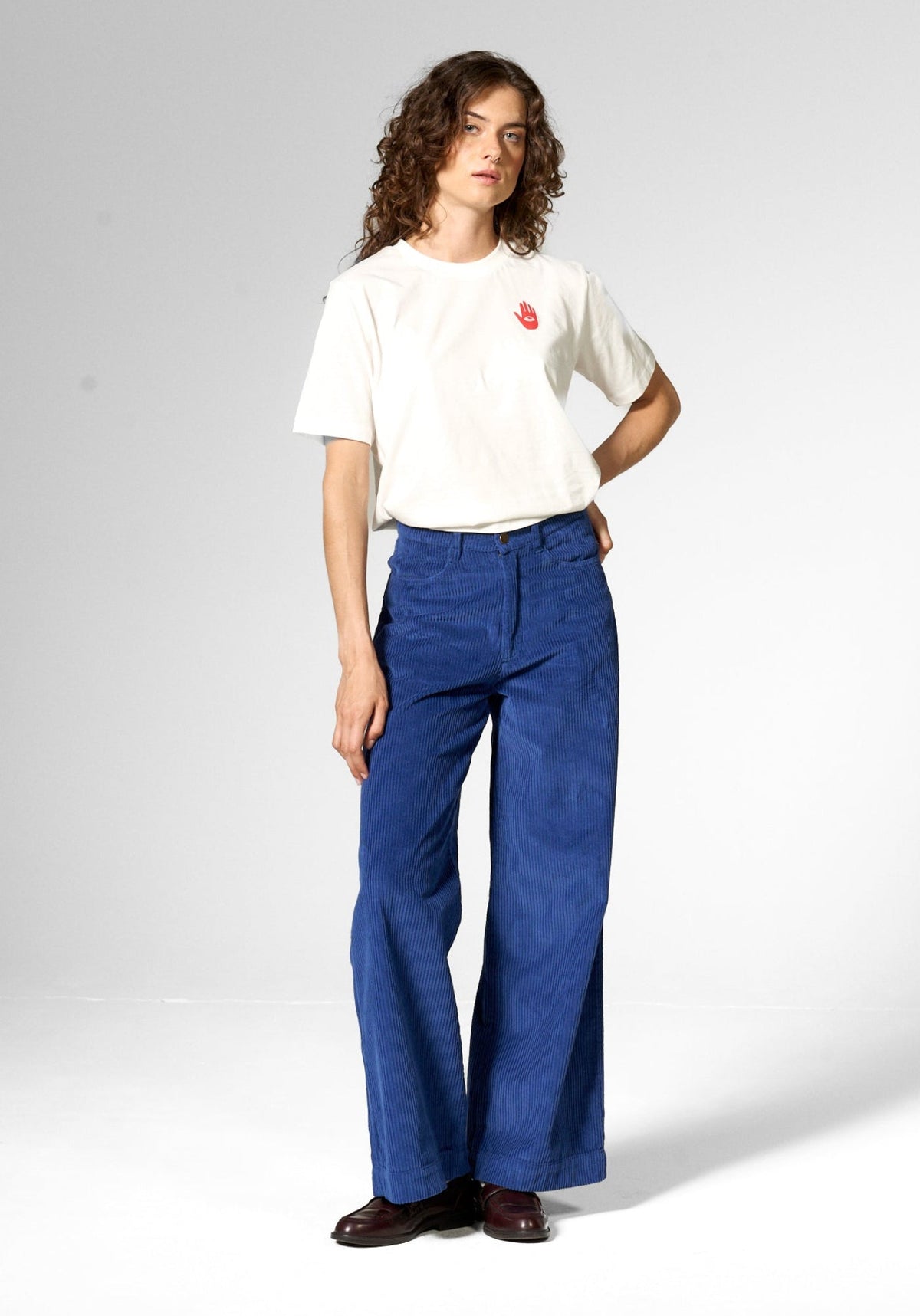 A model stands against a light gray studio background wearing a white t-shirt with a small red graphic on the chest, blue wide-leg pants, and dark brown shoes.