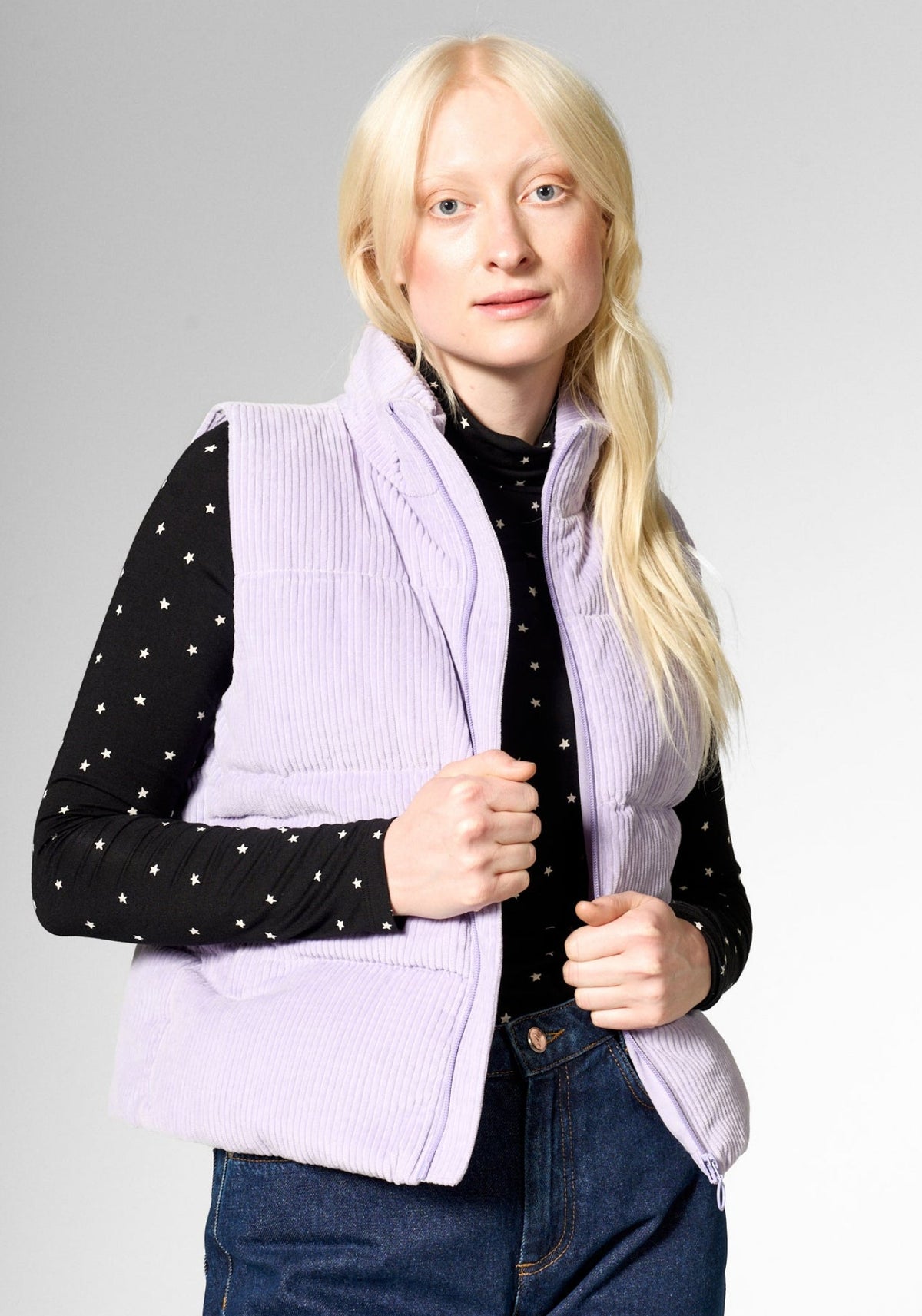 A blonde woman modeling a light purple ribbed corduroy puffer vest over a black long-sleeve top with white polka dots and blue jeans, photographed against a neutral gray studio background.