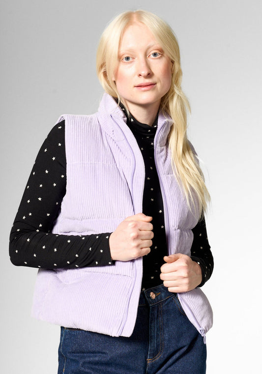 A blonde woman modeling a light purple ribbed corduroy puffer vest over a black long-sleeve top with white polka dots and blue jeans, photographed against a neutral gray studio background.