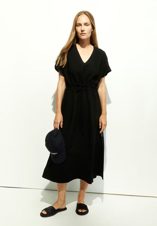 A woman models a black LENZING™ ECOVERO™ and linen blend dress from Armedangels. The dress features a deep V-neckline, short sleeves with a slight flare, and a drawstring waist that creates a cinched silhouette. The hemline reaches just below the knees, and the lightweight, flowy fabric drapes naturally. The person holds a black cap in one hand, wears black slide sandals, and has long, straight hair. They stand against a plain white background with a subtle shadow cast to the side.

