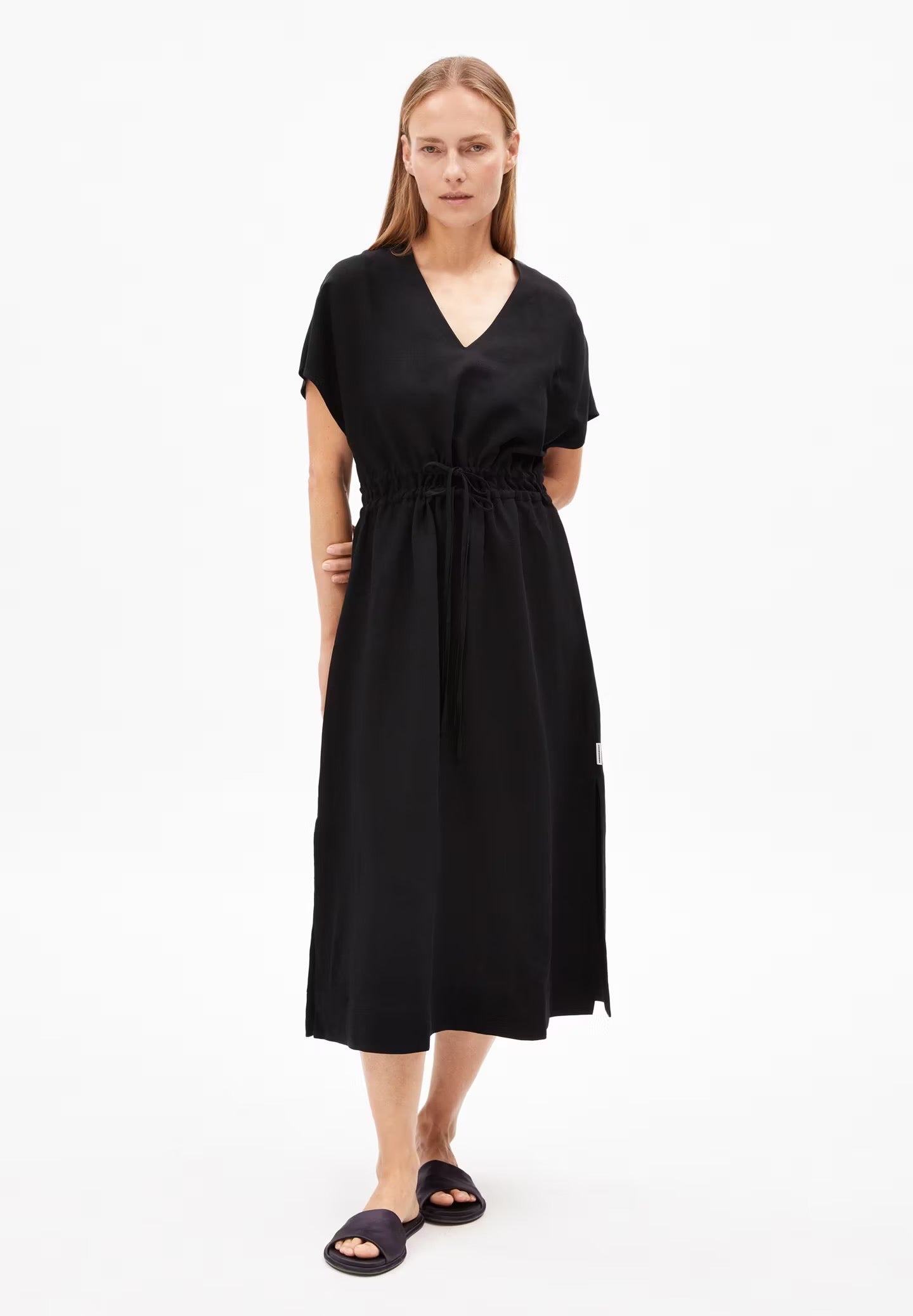 A woman models a black LENZING™ ECOVERO™ and linen blend dress from Armedangels. The dress features a deep V-neckline, short sleeves with a slight flare, and a drawstring waist that creates a cinched silhouette. The hemline reaches just below the knees, and the lightweight, flowy fabric drapes naturally. The person has long, straight hair, wears black slide sandals, and stands with one hand on their hip against a plain white background.

