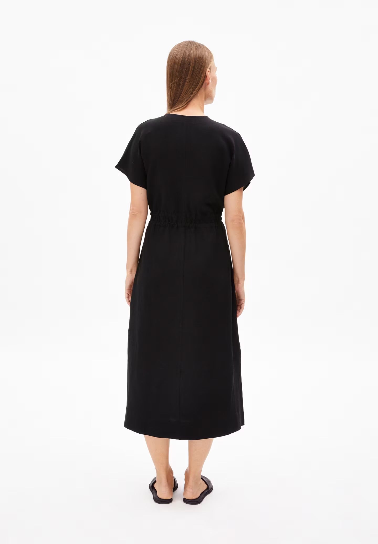 A person models the back view of a black LENZING™ ECOVERO™ and linen blend dress from Armedangels. The dress features a simple, straight silhouette with short, slightly flared sleeves and a cinched waist created by a drawstring tie. The hemline reaches just below the knees, and the lightweight, flowy fabric drapes naturally. The person has long, straight hair and is wearing black slide sandals, standing against a plain white background.

