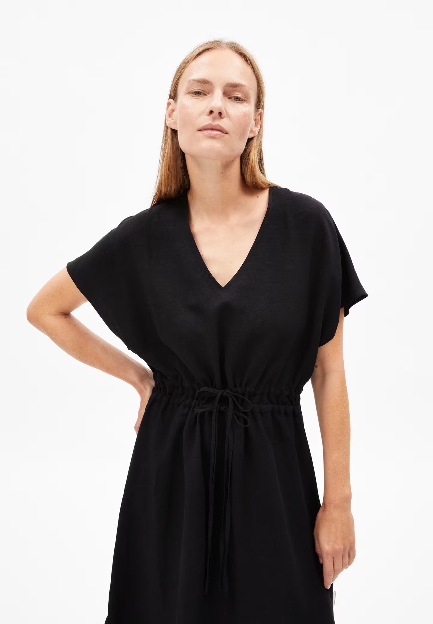 A woman models a black LENZING™ ECOVERO™ and linen blend dress from Armedangels. The dress features a deep V-neckline, short sleeves with a slight flare, and a drawstring waist that creates a cinched silhouette. The fabric appears lightweight and flowy, with a minimalist design, set against a plain white background. The person has long, straight hair and stands with one hand on their hip.

