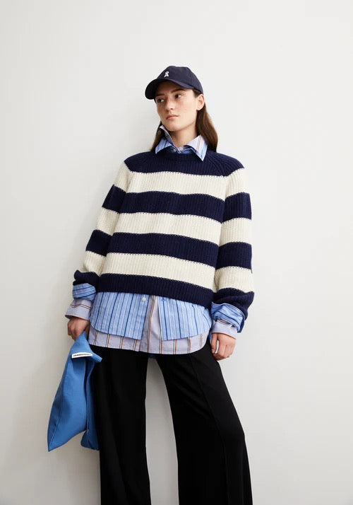 Woman wearing a navy and white striped nautical style jumper from Armedangels made from organic cotton and organic wool,  with layered shirts and black pants against a plain background