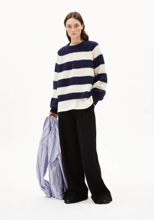 Woman wearing a navy and white striped nautical style chunky knit jumper from Armedangels made from organic cotton and organic wool, with black pants on a white background