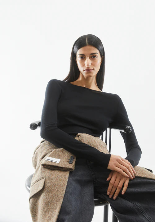 Woman sitting on a chair wearing a black long sleeved wool and Tencel blend top by Armedangels and beige coat against a white background