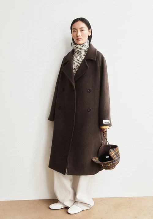 Woman wearing a brown woolen coat with a plaid bag against a white wall