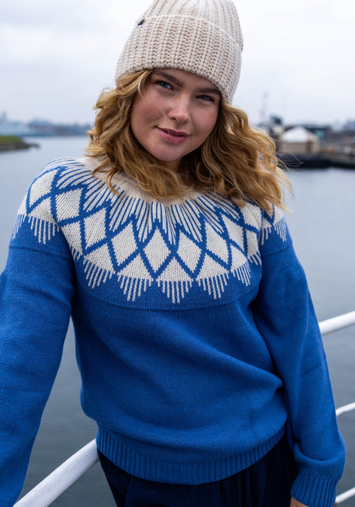 A woman wearing a blue and cream knit jumper by Danefae with a geometric yoke pattern, a cream knit beanie, and dark trousers, standing outdoors near a railing with water and a distant harbor in the background.