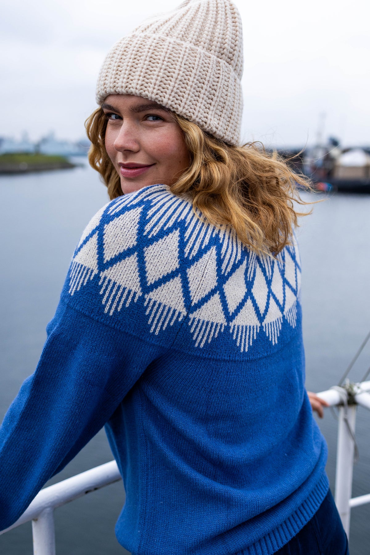 A woman on a boat wearing a blue and white patterned knit jumper by Danefae and a cream-colored knit beanie, looking back over her shoulder.