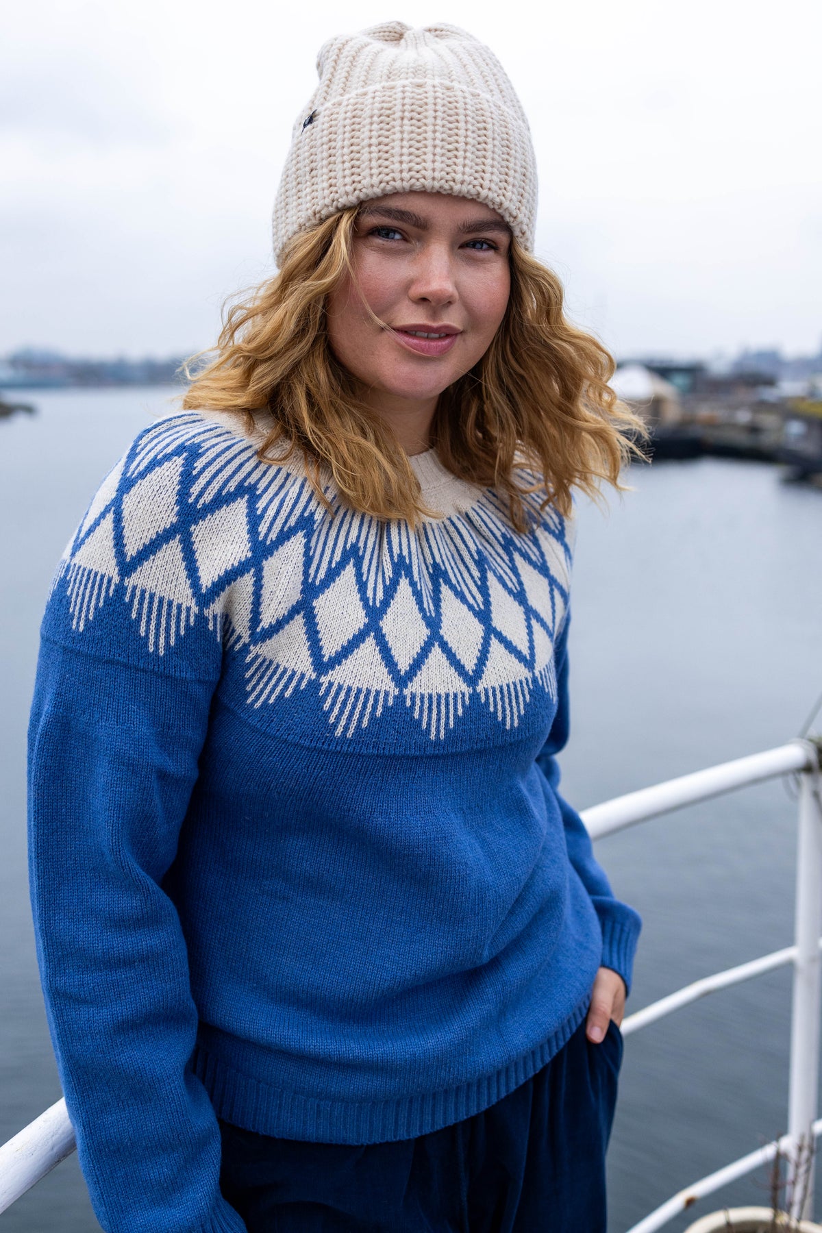 A woman wearing a blue and cream Fair Isle knit jumper by Danefae and a cream beanie, standing outdoors near a body of water with a railing in the foreground.