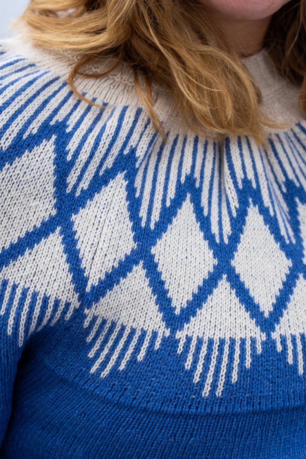 Close-up of a person wearing a blue and off-white knitted yoke jumper by Danefae with a geometric diamond pattern.
