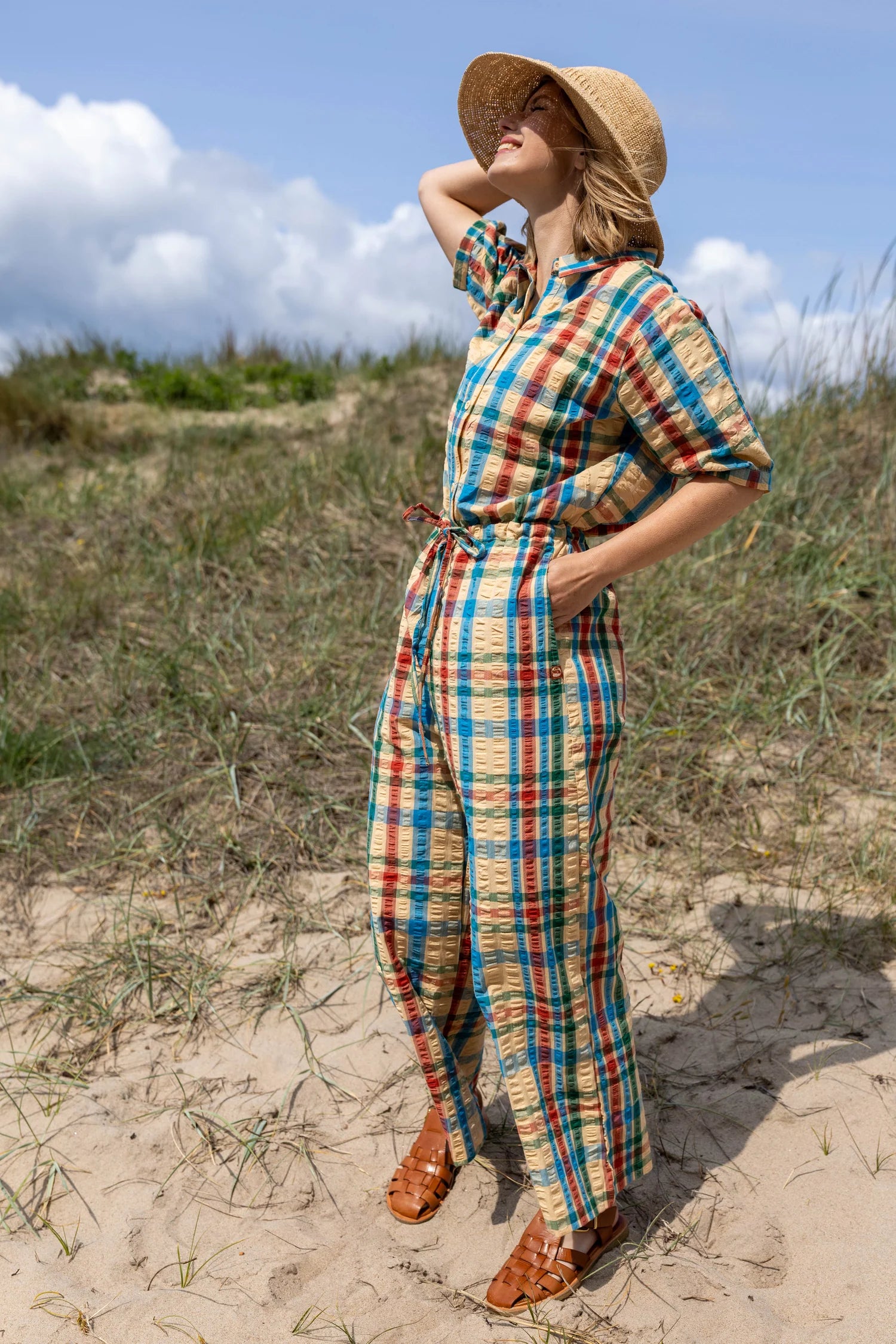 Woman in a plaid jumpsuit and straw hat standing on sandy dunes with a blue sky.