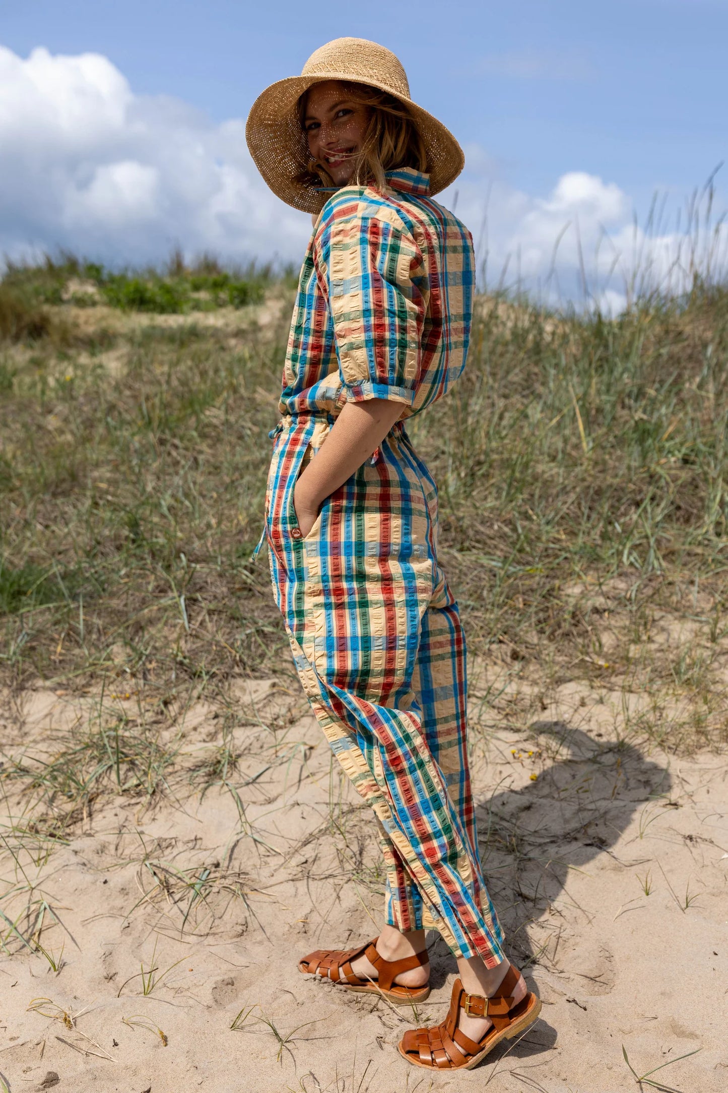 Woman in a plaid jumpsuit standing on sandy dunes with grass and blue sky in the background