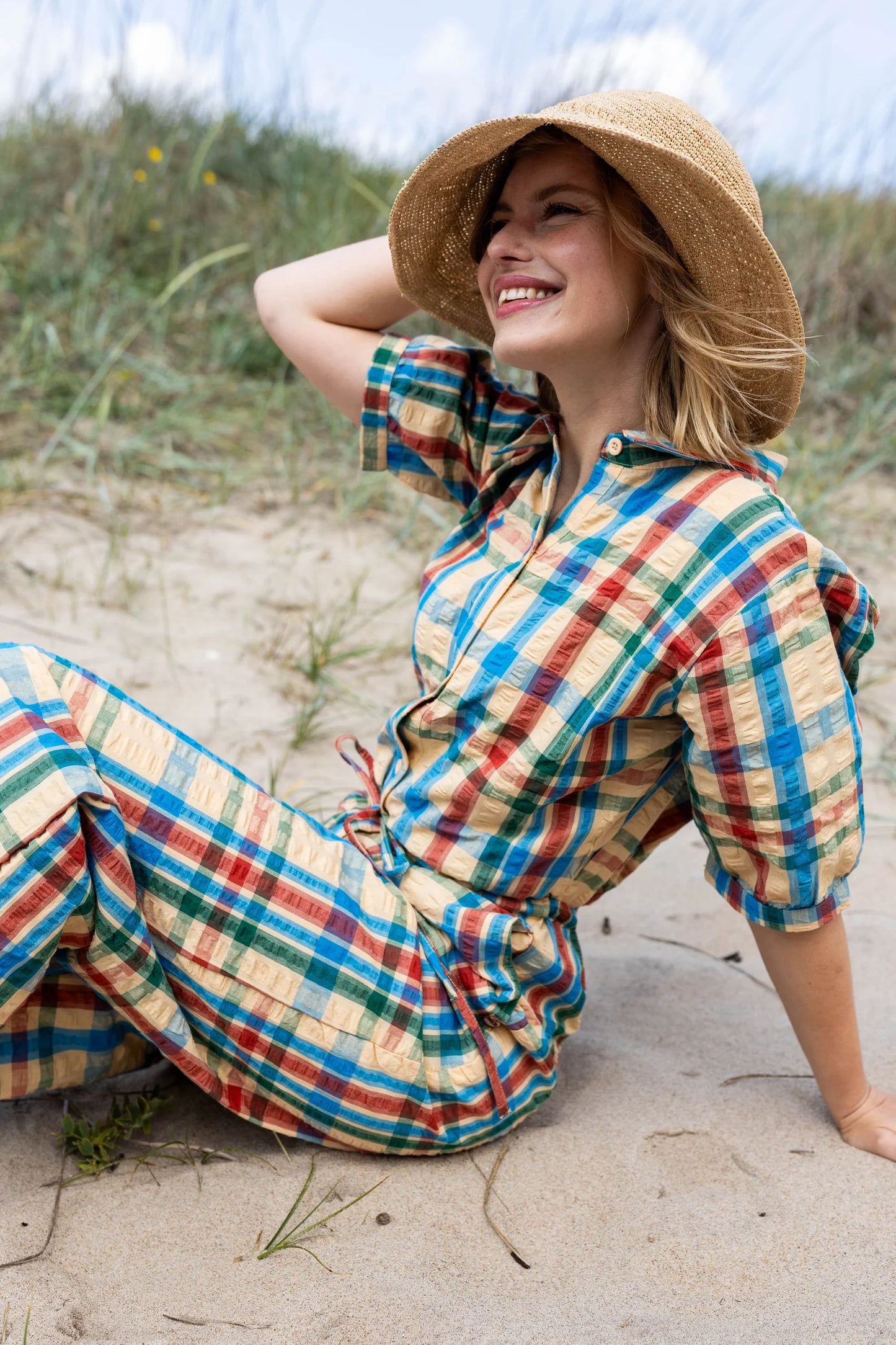 Woman in a colourful plaid jumpsuit and sun hat sitting on sand with grass and sky in the background
