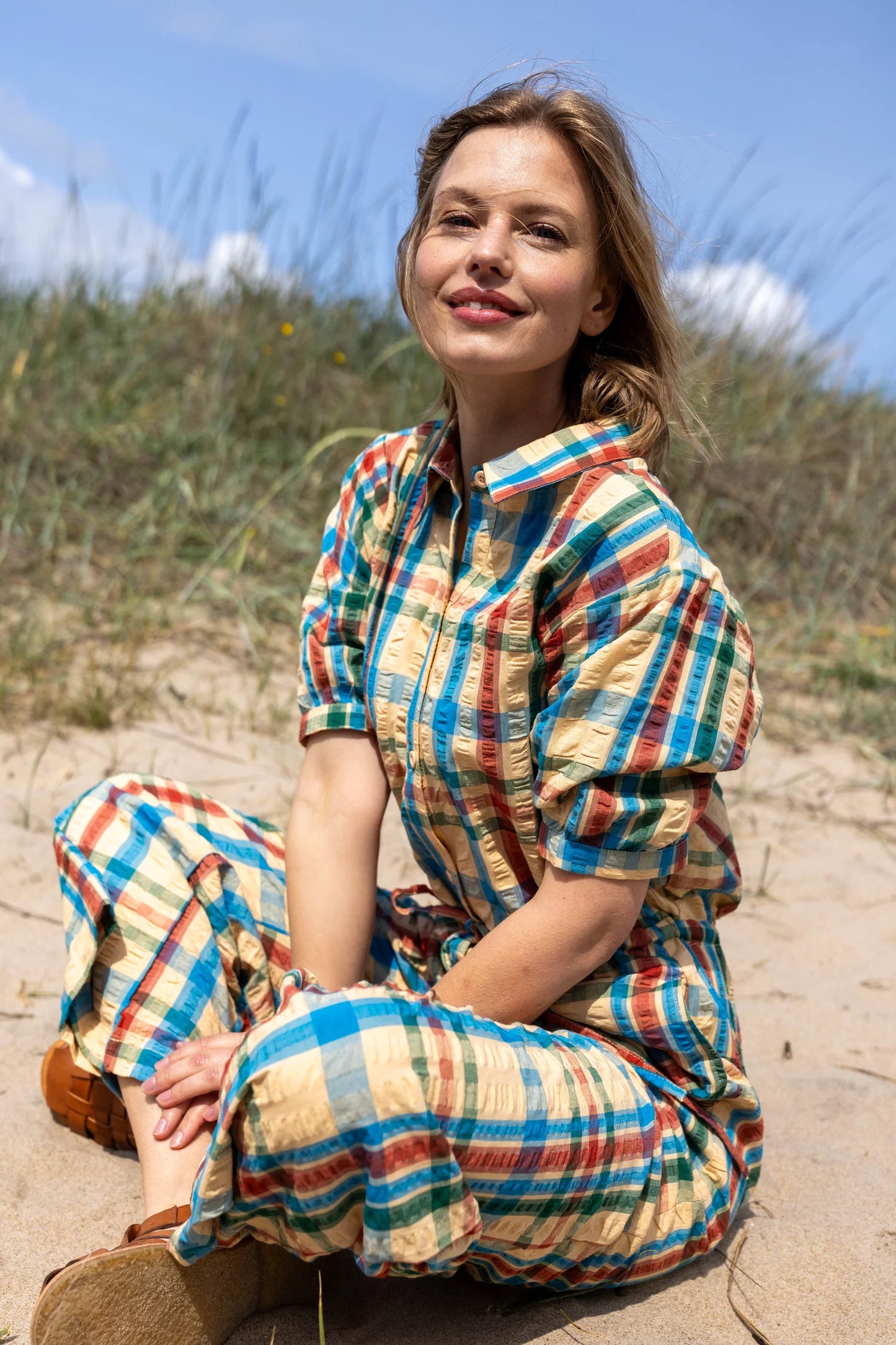 Woman sitting on a sandy beach wearing a colourful plaid jumpsuit.