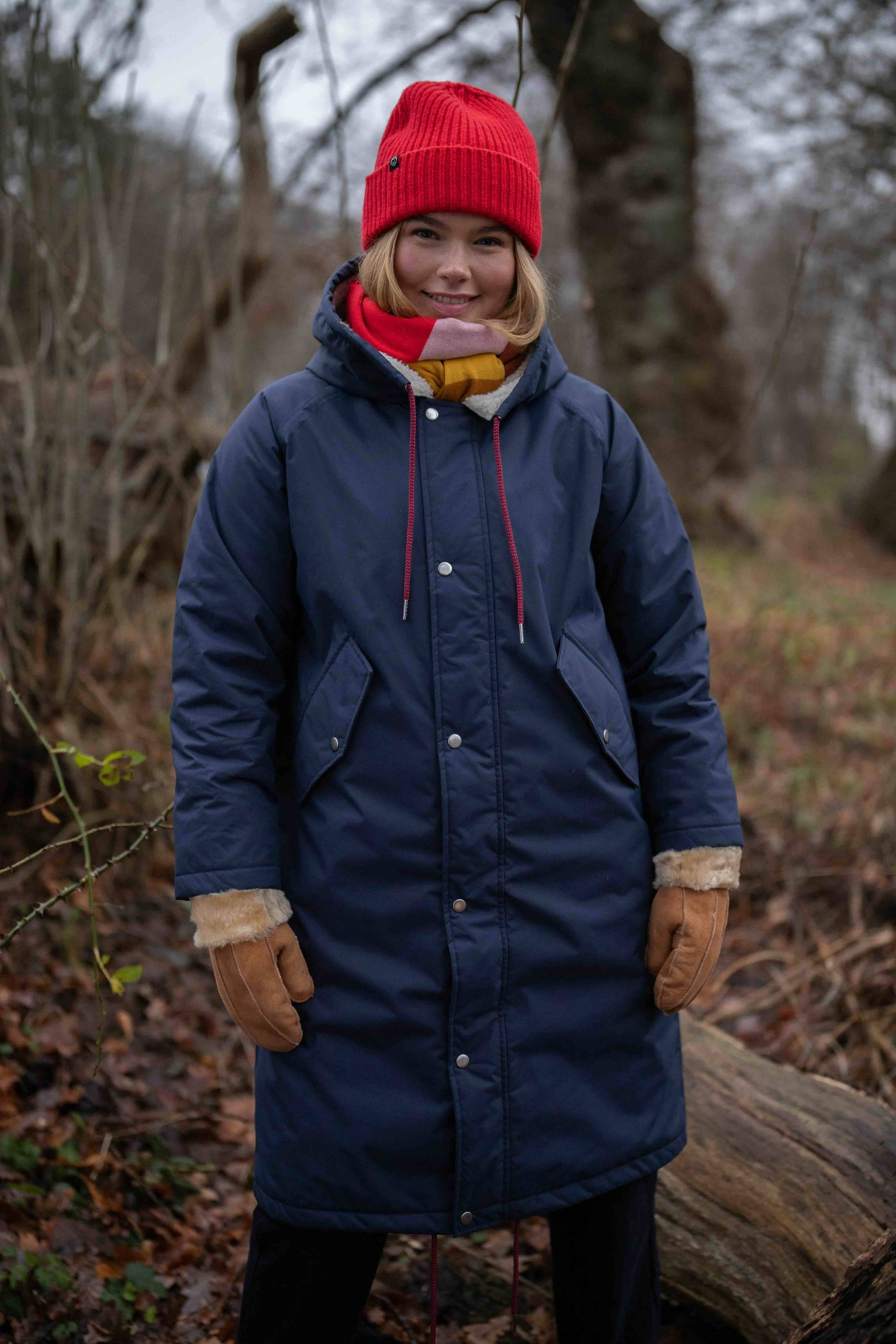 Woman wearing the Danecoast waterproof winter coat from Danefae Copenhagen in navy blue, a red beanie, and brown gloves standing outdoors in a forest.