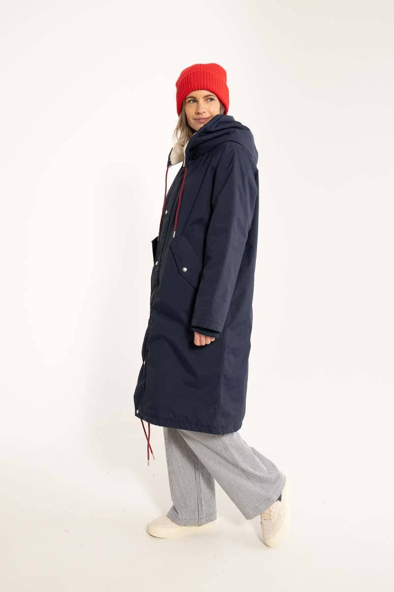 Woman wearing a long navy coat, the waterproof Danecoastred winter parka from Danefae Copenhagen, paired with a beanie, and gray pants on a white backgroun