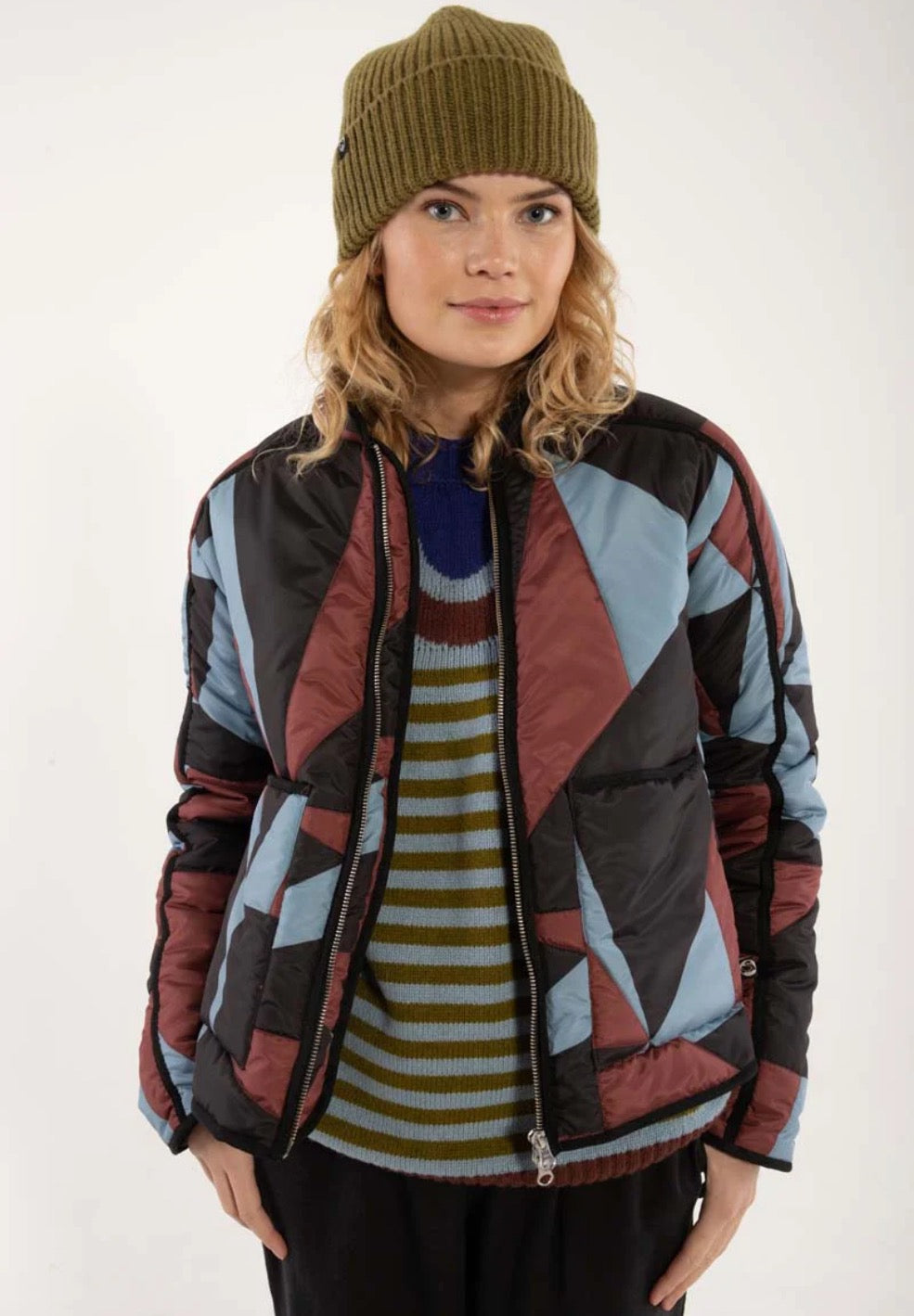 Woman wearing a thermal jacket from Danefae, featuring a geometric pattern in ice blue, wine red and black, and green beanie on a white background