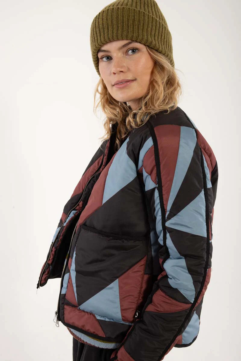 Woman wearing a thermal jacket from Danefae, featuring a geometric pattern in ice blue, wine red and black, and green beanie on a white background