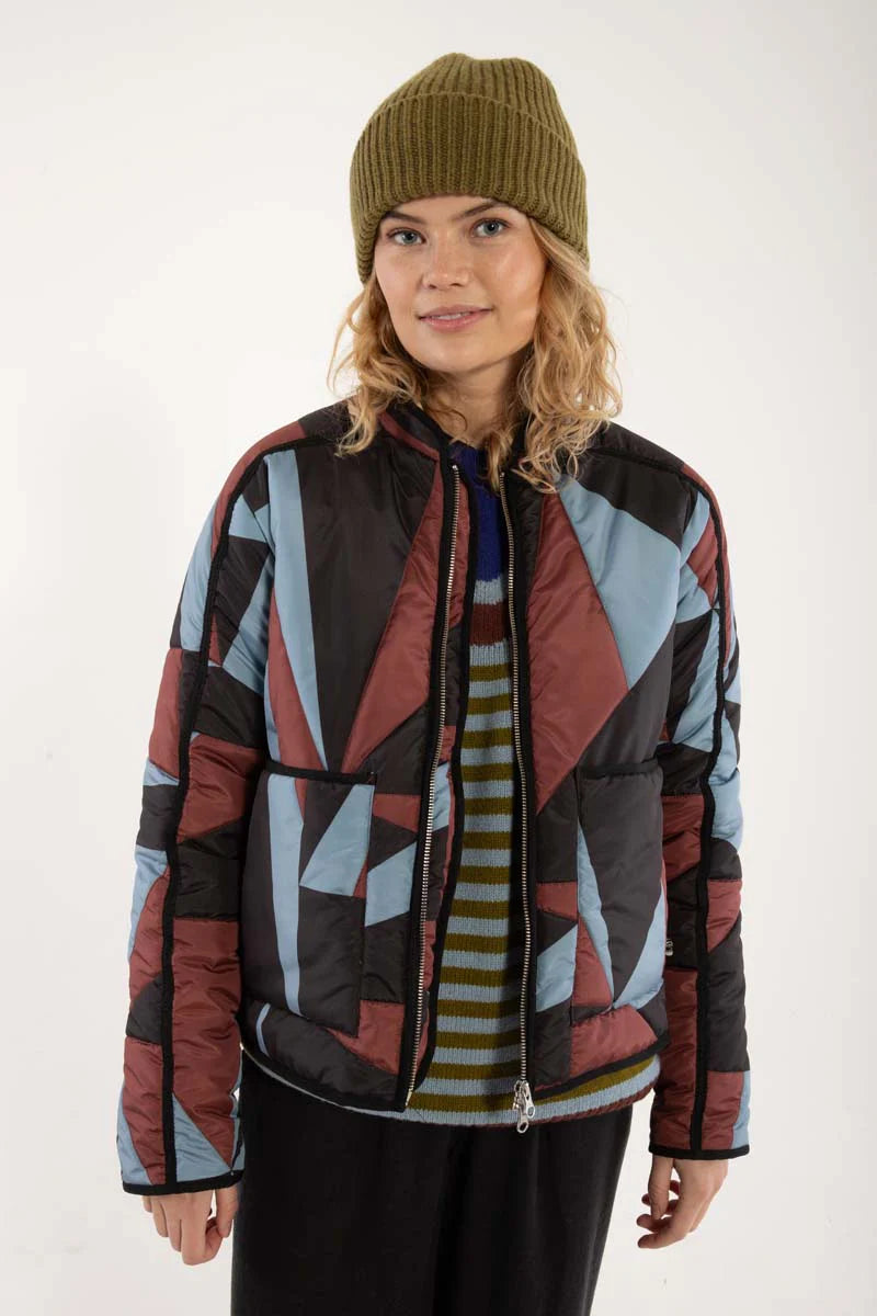 Woman wearing a thermal jacket from Danefae, featuring a geometric pattern in ice blue, wine red and black, and green beanie on a white background