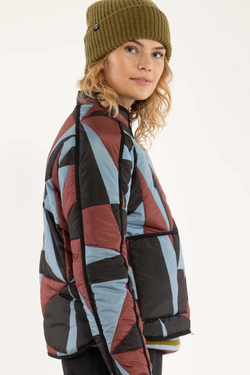 Woman wearing a thermal jacket from Danefae, featuring a geometric pattern in ice blue, wine red and black, and green beanie on a white background