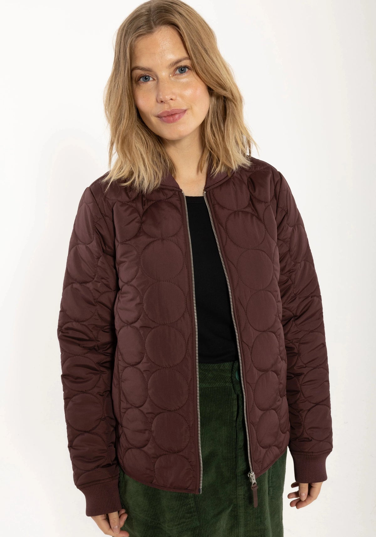 A woman wearing a dark red quilted jacket from Danefae with a circular bubble pattern, styled over a black top and green skirt, against a plain white background.