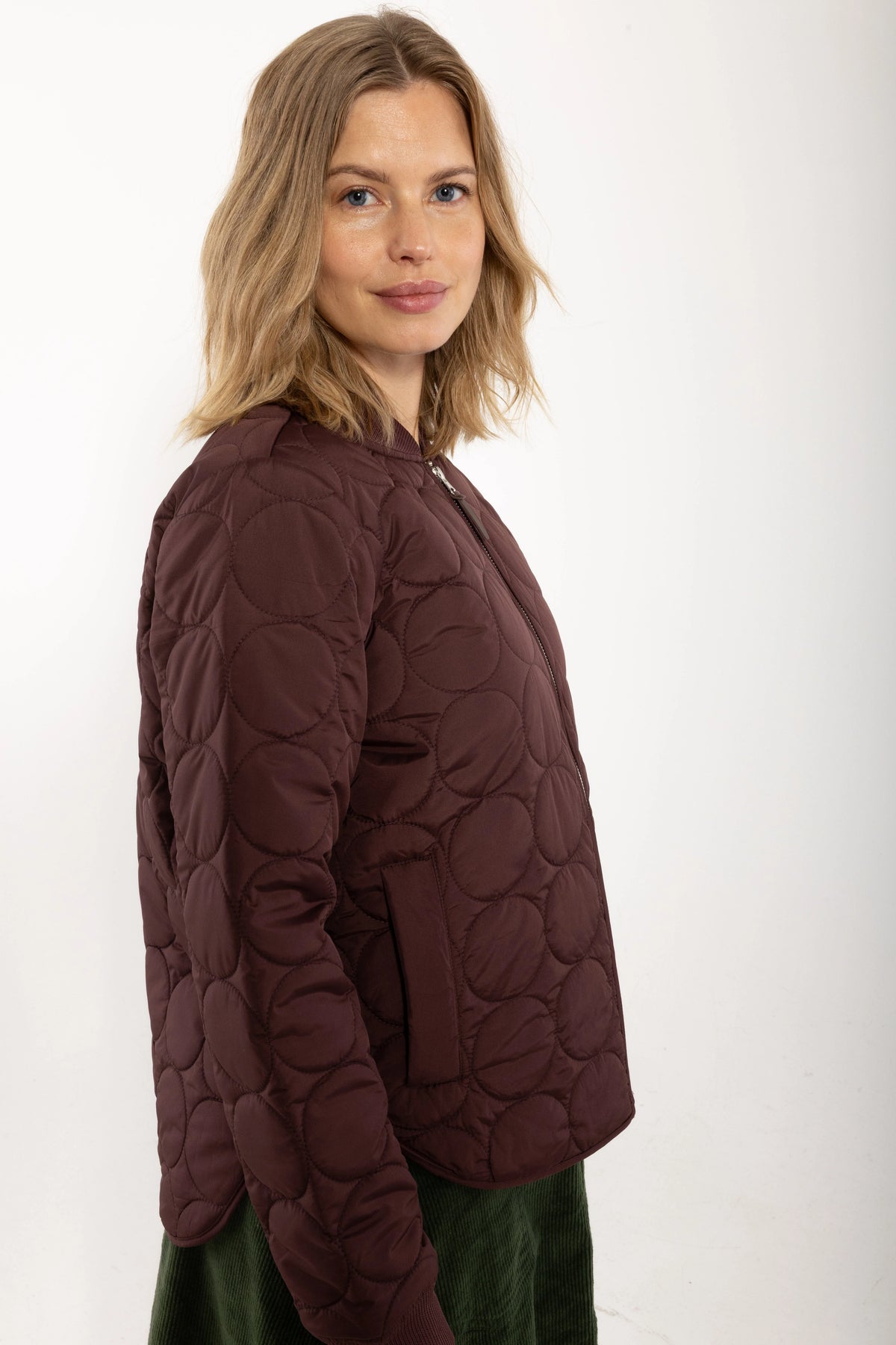 A woman wearing a dark red quilted jacket by Danefae with a circular stitched pattern, shown from a side-back angle against a plain white studio background.